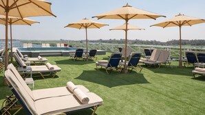 A rooftop lounge area with sun loungers and umbrellas, overlooking a body of water and cityscape.
