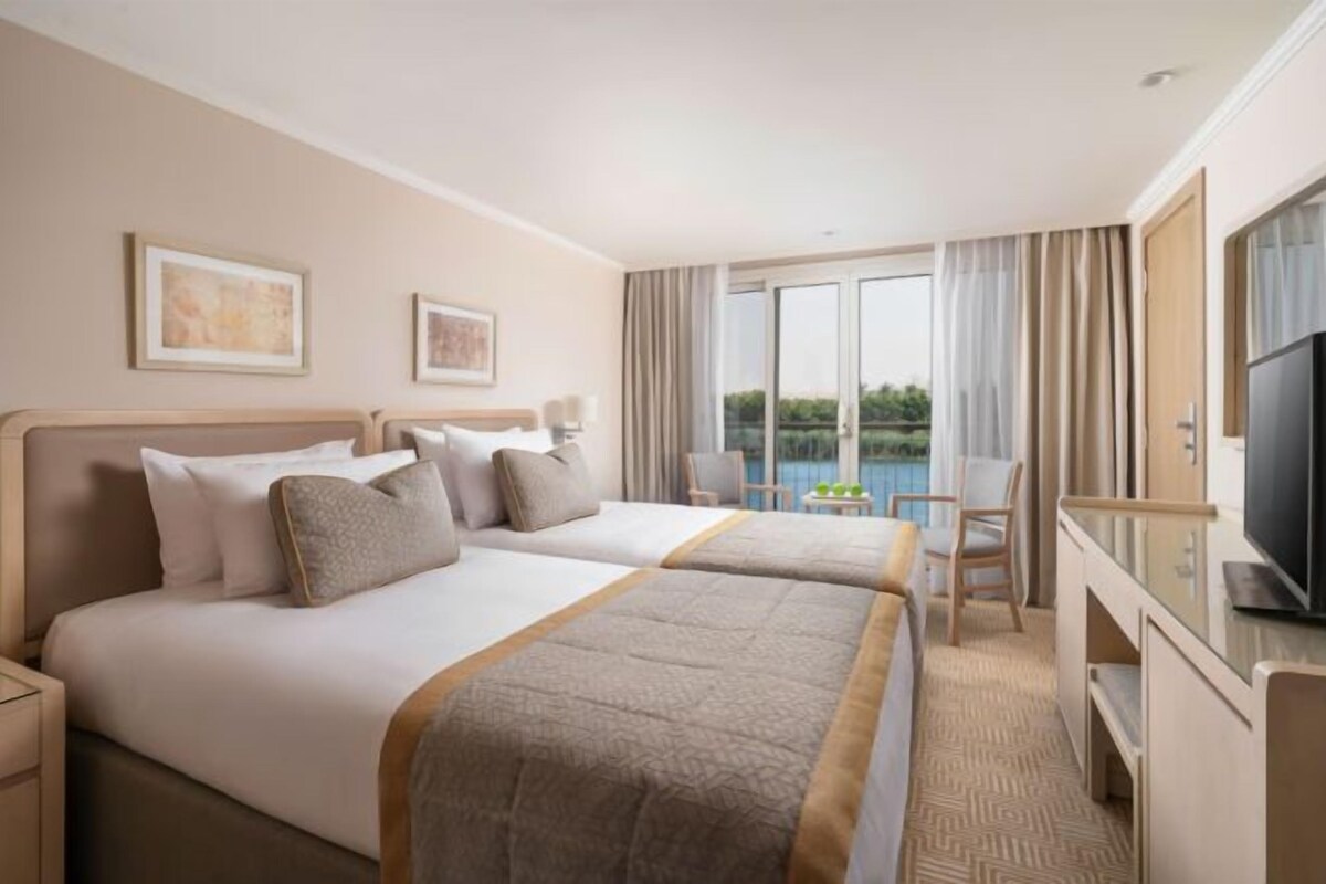 A hotel room with two beds, a desk, a TV, and a balcony with a view.