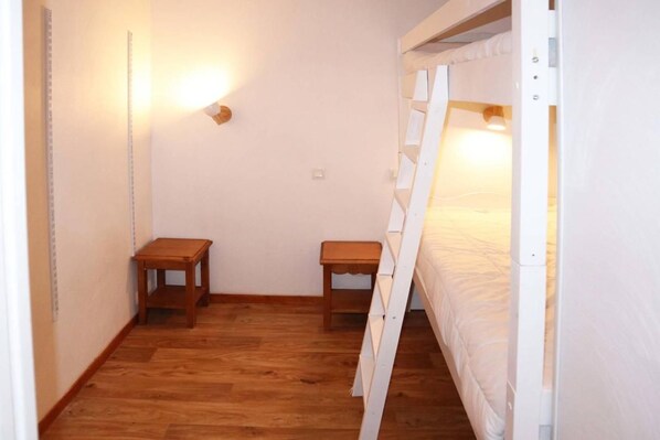 A small, narrow room with a bunk bed, two wooden nightstands, and a wall-mounted light fixture.
