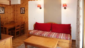 A cozy room with a wooden dining table, chairs, a sofa with red cushions, and two wall-mounted lamps.