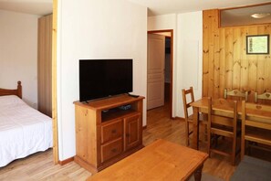 A room with a bed, a TV on a wooden stand, a dining table with chairs, and a wooden wall panel.