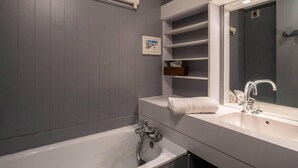 A modern bathroom with a bathtub, two sinks, and a large mirror. There are shelves above the sinks and a small framed picture on the wall.
