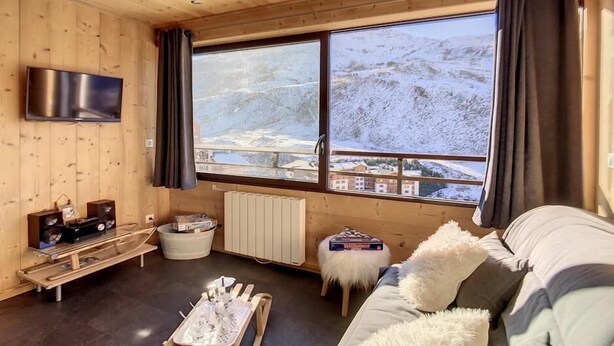 Interior acogedor de una cabaña con un amplio ventanal que ofrece vistas a una montaña nevada, un televisor de pantalla plana montado en la pared y una zona de estar cómoda con un sofá y una mesita.