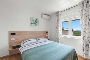 Une chambre avec une tête de lit en bois, un lit avec une literie à rayures, un climatiseur fixé au mur, un tableau encadré et une fenêtre avec des rideaux à motifs.