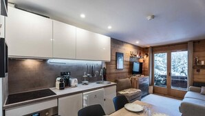 A modern kitchen with white cabinets, a wooden dining area with a table set for two, and a large window with a snowy view. - 90m from ski slopes, floor -2, balcony, tv, 36m², Méribel-Mottaret (Les Allues)