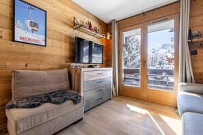 A cozy room with a sofa, a TV, a wooden wall, a shelf with decorations, and a large window with a view of snow-covered trees. - 350m from shops, floor -2, balcony, tv, 36m², Méribel-Mottaret (Les Allues)