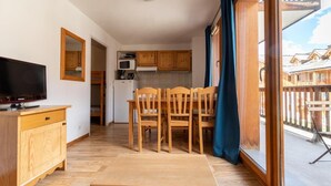 A compact room with a wooden dining table and chairs, a television on a wooden cabinet, a kitchenette with wooden cabinets, and a balcony with a wooden railing.