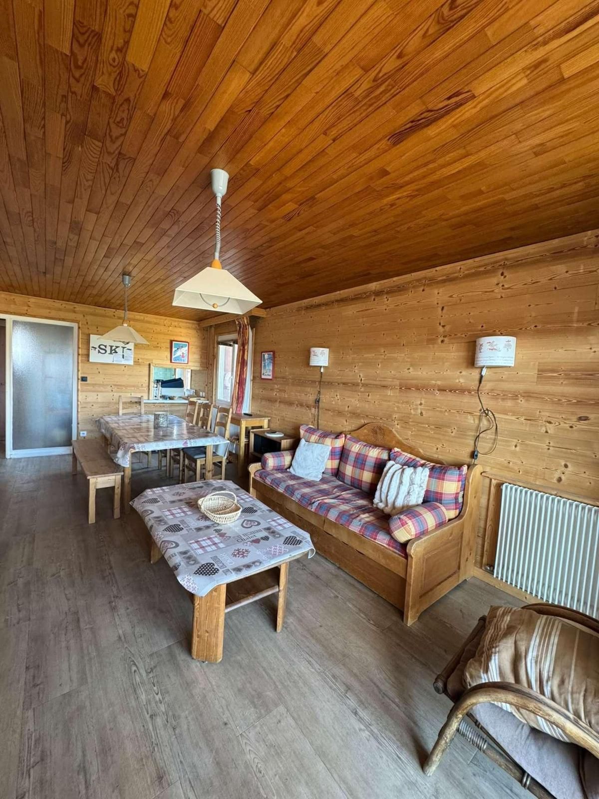 A cozy cabin interior with a wooden ceiling, a sofa with plaid cushions, a dining table with chairs, and a bench.