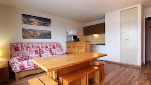 Un espacio habitable compacto con una mesa de comedor de madera, bancos, un sofá con cojines estampados, una zona de cocina con armarios y electrodomésticos, y dos cuadros en la pared.