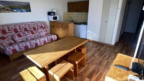 Un espacio habitable pequeño con una mesa de comedor de madera, bancos, un sofá estampado, una kitchenette y una puerta que da a otra habitación.