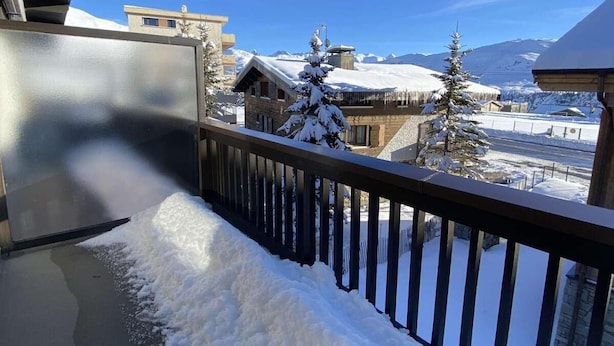 Un balcón con barandilla de metal que da a un paisaje nevado con edificios y montañas al fondo.
