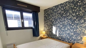 Habitación individual con una cama de madera, una ventana con cortinas, un radiador y una mesita de noche con lámpara, todo sobre un fondo de papel tapiz floral.