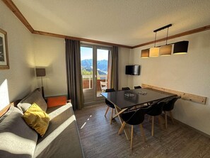 A cozy living room with a sofa, a dining table with chairs, a TV mounted on the wall, and a balcony with a view of the mountains. - 130m from ski slopes, 7th floor, swimming pool, sauna, balcony, tv, ski locker, 59m², Alpe d'Huez (L'Alpe d'Huez)