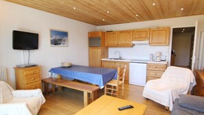 Et hyggeligt værelse med trælofter, et spisebord dækket af en blå dug, et fjernsyn på et træskab, et køkken med træskabe og en hvid sofa.