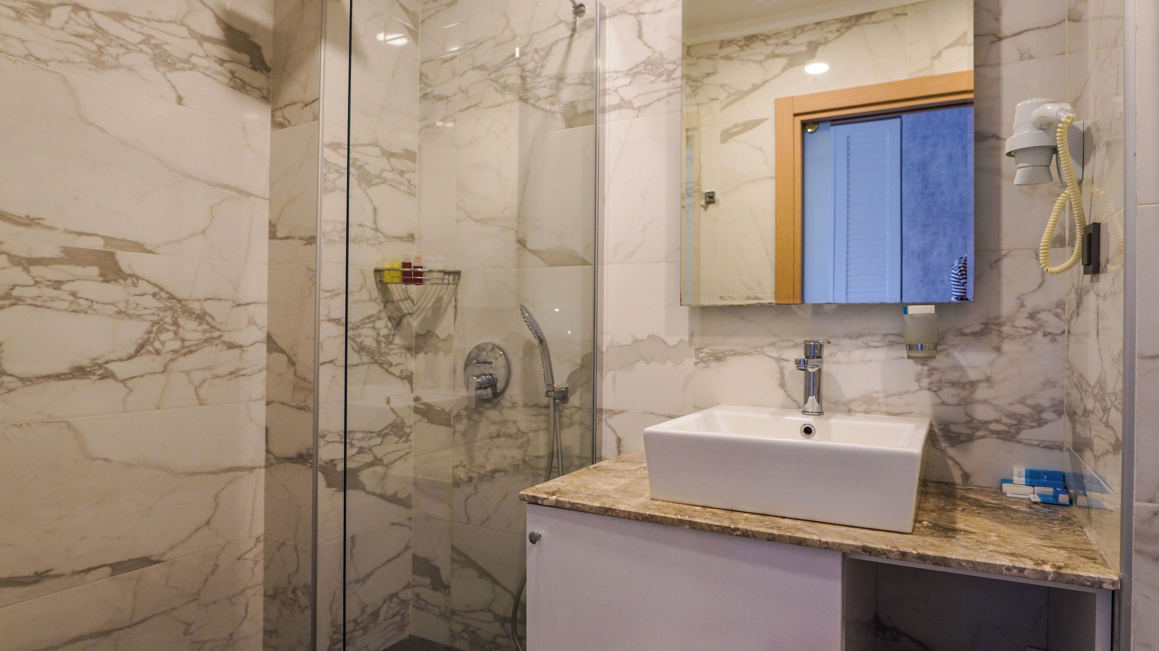 Un bagno con parete in marmo, box doccia in vetro, lavabo con specchio sopra e telefono fissato alla parete.