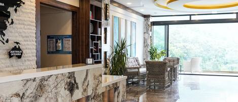 A modern lobby with a marble reception desk, wooden paneling, and a view of greenery outside.