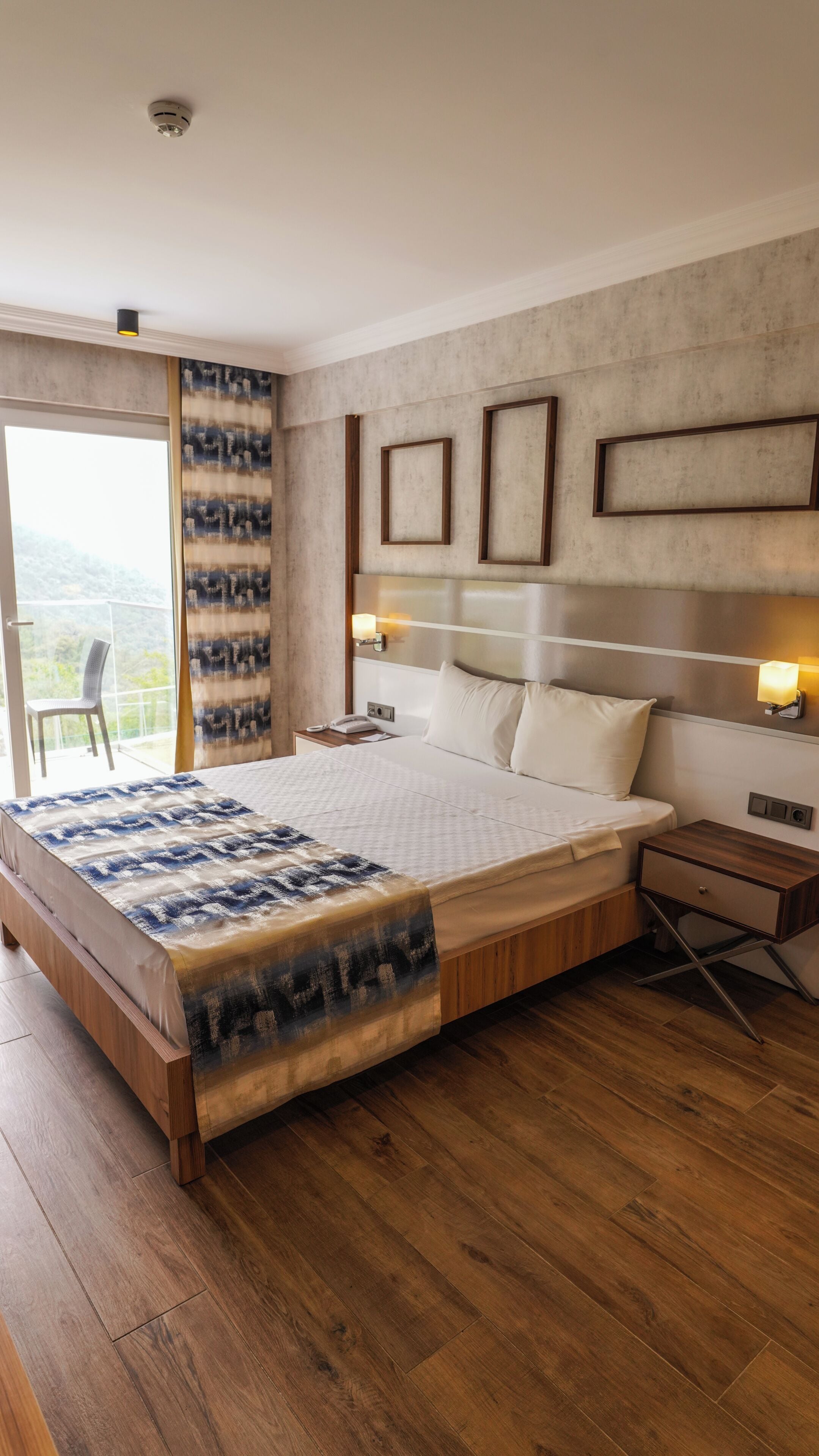 A modern hotel room with a large bed, wooden floor, a balcony with a chair, and a view of the outdoors.