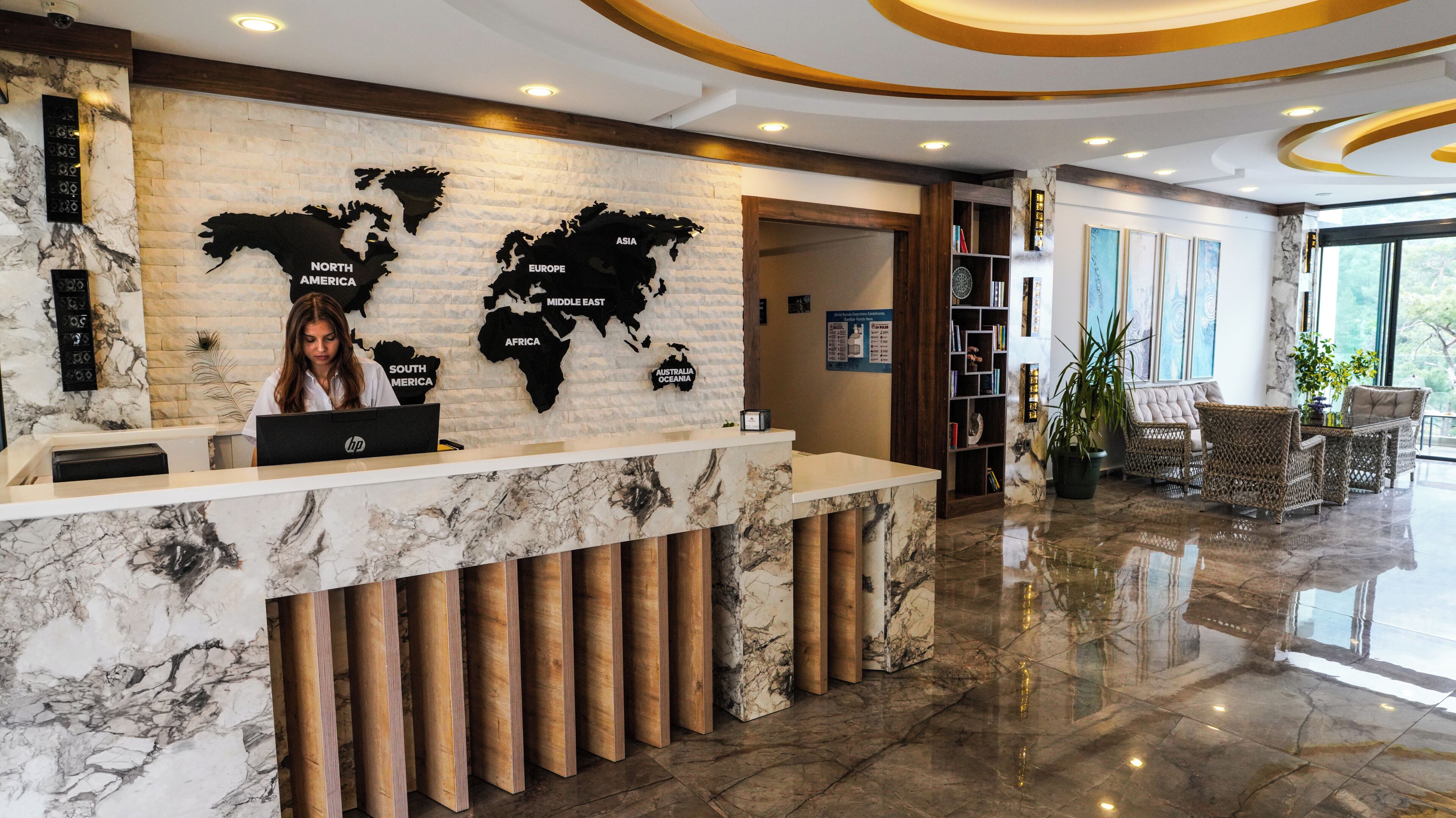 Mostrador de recepción de hotel con un mapa mundial, mostrador para recibir a los huéspedes, zona de estar con mobiliario de mimbre y un amplio ventanal.