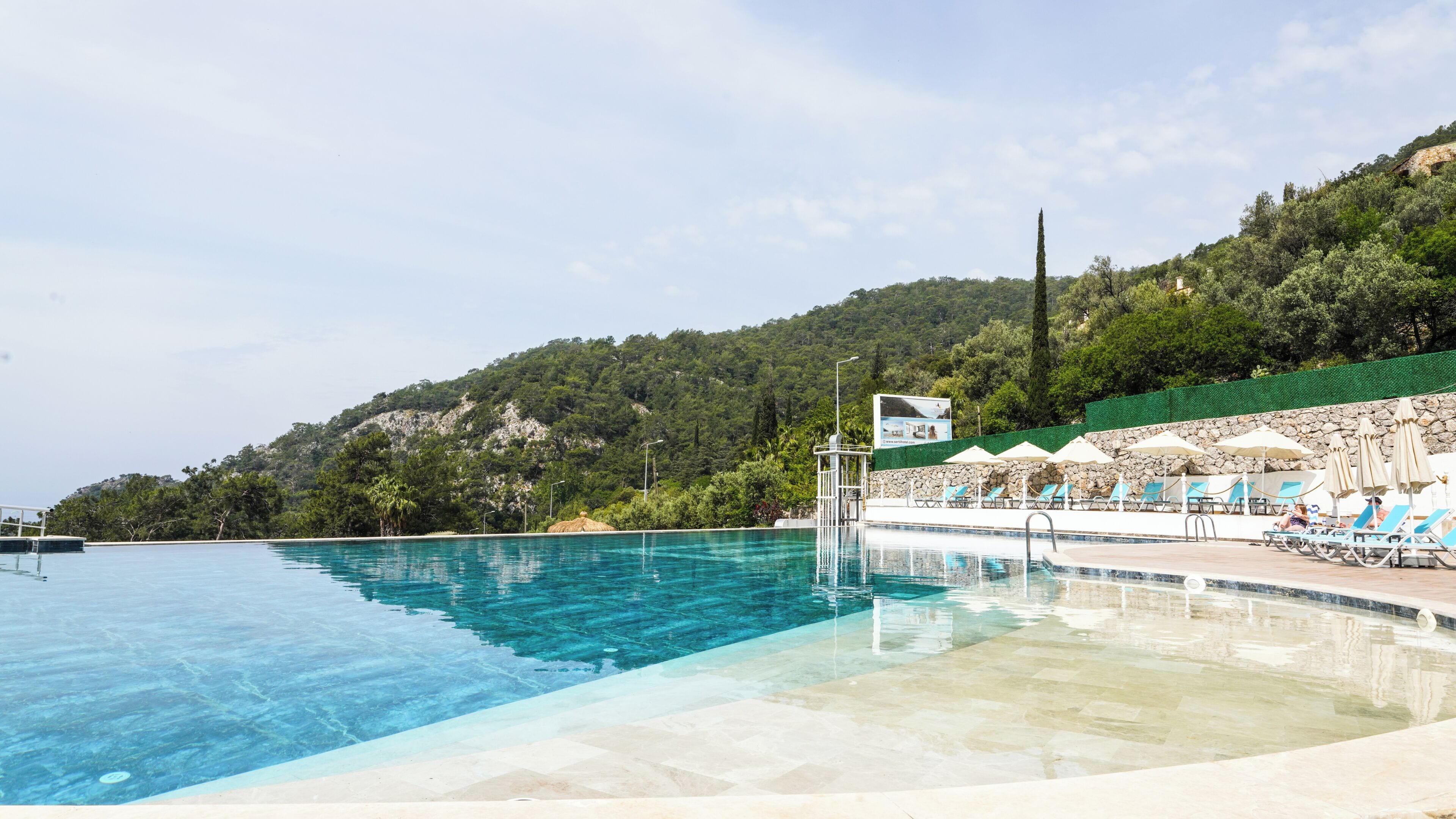 A swimming pool with clear water, surrounded by lounge chairs and umbrellas, set against a backdrop of a green hillside.