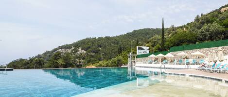 A swimming pool with clear water, surrounded by lounge chairs and umbrellas, set against a backdrop of a green hillside.