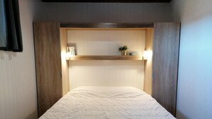 Un lit simple avec une couette blanche, dans une pièce aux murs lambrés et agrémentée d’une étagère où sont disposés des objets décoratifs.