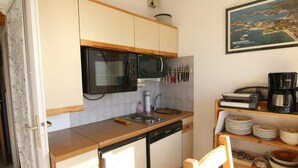 Een kleine keuken met een magnetron, spoelbak en vaatwasser. Er is een houten plank met servies en een lijst met een foto aan de muur.