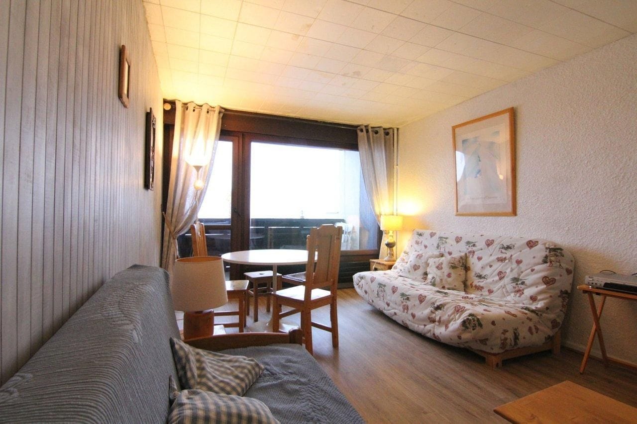 A cozy living room with a sofa, a patterned couch, a dining table with chairs, and a window with curtains.
