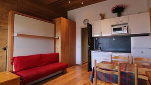 Cocina compacta con microondas, refrigerador y lavamanos. Un sofá rojo y una mesa de comedor de madera con sillas se encuentran en la zona de estar.