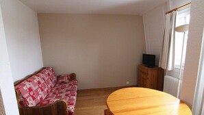 Una habitación con piso de madera, una mesa redonda de madera, un sofá con estampado rojo y blanco, y un televisor sobre un mueble de madera.