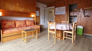 Une pièce chaleureuse, meublée de meubles en bois, notamment un canapé, une petite table et une table à manger avec des chaises. On y trouve une porte qui donne sur une autre pièce, ainsi qu'une poubelle verte.