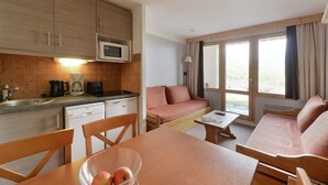 Eine kompakte Küche mit Mikrowelle, Kaffeemaschine und Spüle. Ein Esstisch mit Stühlen und eine Sitzecke bieten Blick ins Freie.