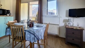 En matplats med ett bord för fyra personer, ett köksområde med mikrovågsugn och kaffebryggare, ett fönster med utsikt över bergen och en tv på väggen.