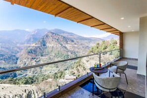 A balcony with a view of a mountainous landscape, a table with chairs, and a glass railing. - Regenta Resort Madhuganga Ukhimath (Ukhimath)