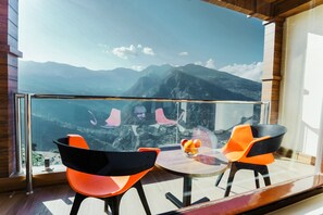 A modern room with a large window offering a mountain view. There are two orange chairs and a small table with a fruit bowl. - Regenta Resort Madhuganga Ukhimath (Ukhimath)