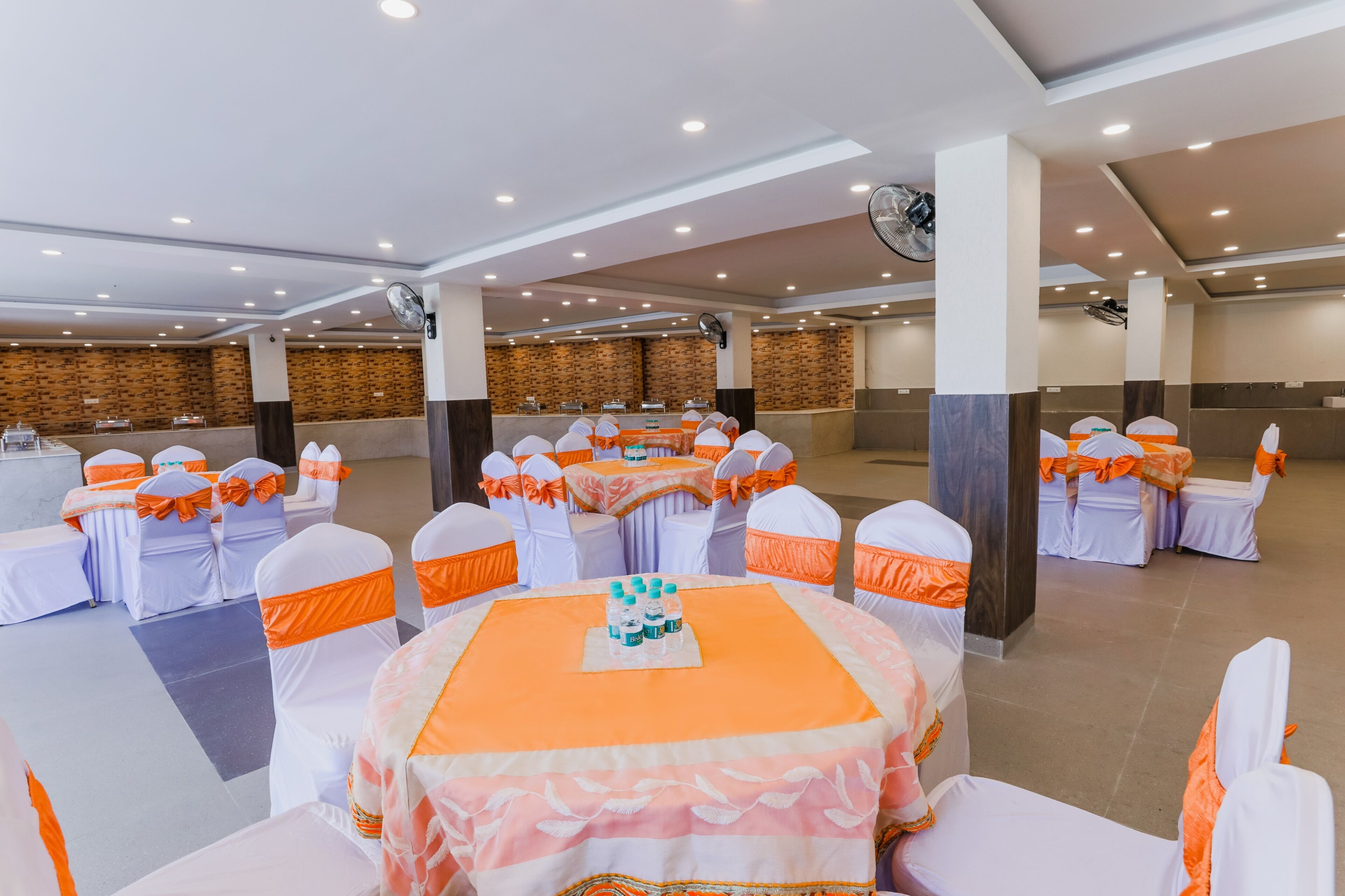Un salón de banquetes con mesas redondas, sillas blancas y anaranjadas, y una pared de ladrillo al fondo.