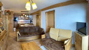Una sala de estar acogedora con un sofá, una mesa de centro y un televisor. Se observa una cocina al fondo con armarios de madera y un microondas.