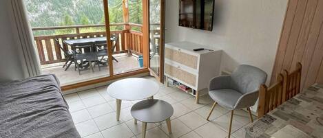 A room with a bed, two small tables, a sofa, a TV, and a balcony with a table and chairs.