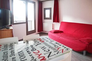A room with a red sofa, a television on a wooden stand, and a window with curtains.