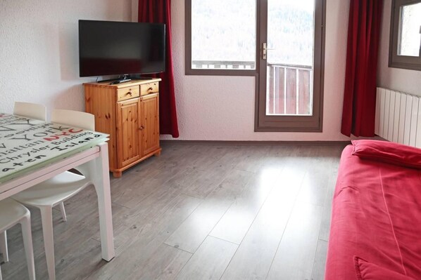 A room with a television on a wooden cabinet, a table with papers, a red couch, and a window with curtains.