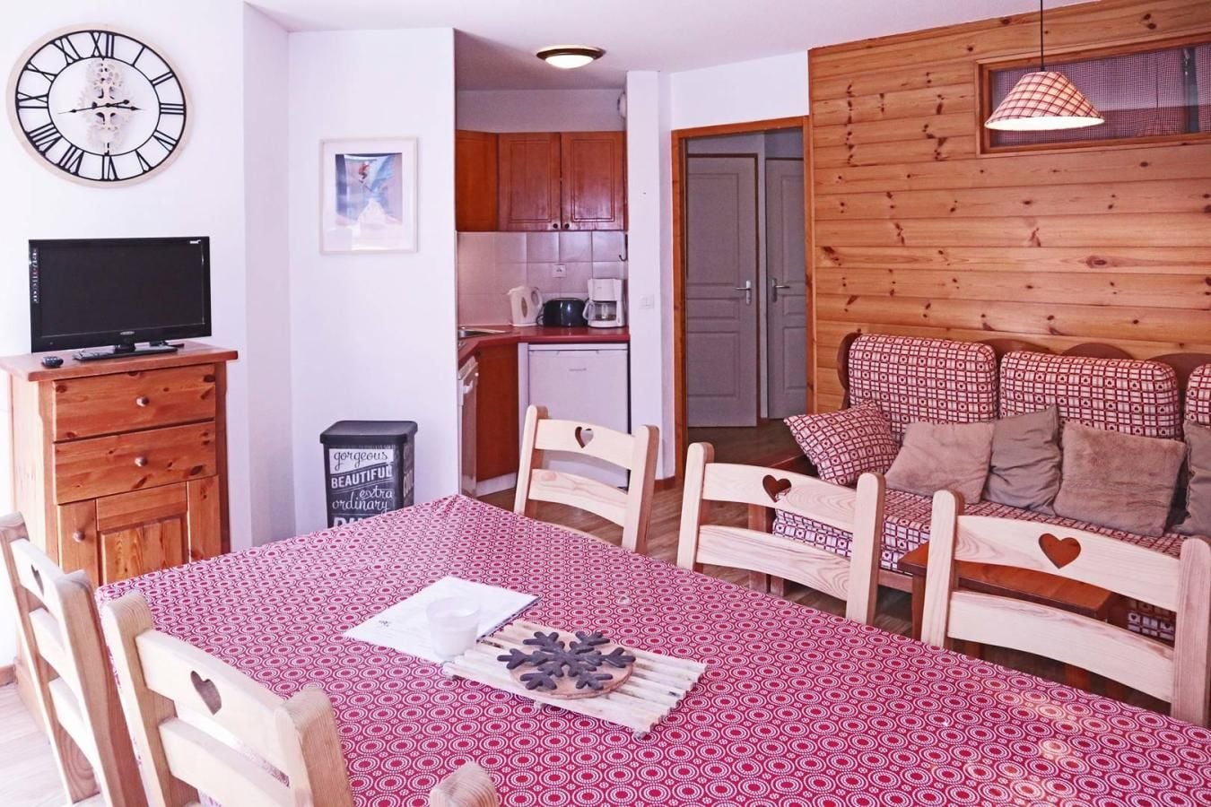 Un accogliente soggiorno con un tavolo da pranzo, sedie di legno, un divano con cuscini decorati, una televisione e una cucina sullo sfondo.