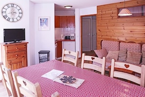 A cozy living room with a dining table, wooden chairs, a sofa with patterned cushions, a television, and a kitchen in the background.