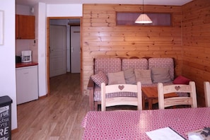 A cozy room with a wooden wall, a patterned sofa, a small table, and a kitchenette area. - Ski-in/ski-out & foot of the slopes, 2nd floor, view ski slopes, swimming pool, sauna, balcony, tv (Les Orres)