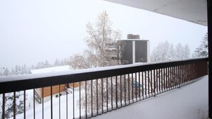 Un balcon enneigé avec une rambarde en métal, un bâtiment avec un réservoir d’eau en arrière-plan et des arbres couverts de neige.