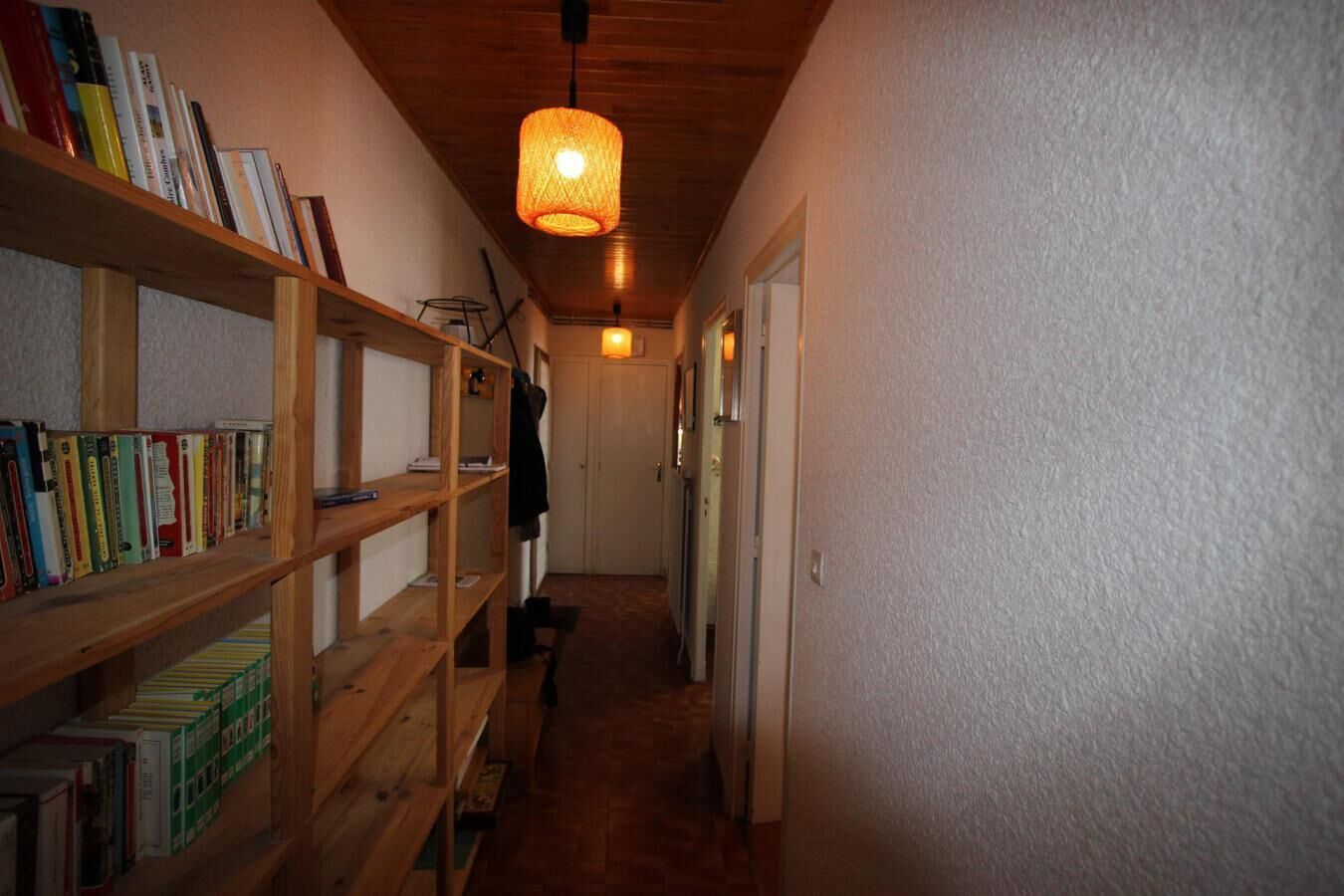 Een smalle gang met houten boekenkasten aan één kant en een hanglamp in het midden.