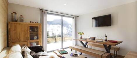 A cozy living room with a sofa, a wooden cabinet, a dining table with benches, and a TV mounted on the wall.