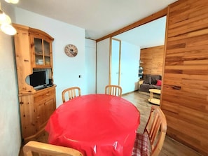 A dining area with a round table covered in a red tablecloth, wooden chairs, a wooden cabinet with a television, and a clock on the wall.