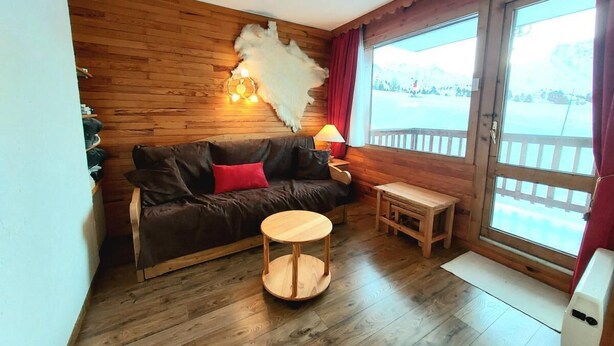 Un cuarto acogedor con piso de madera, un sofá de cuero marrón con cojines rojos, una mesa de madera redonda y un ventanal con vistas a un paisaje nevado.