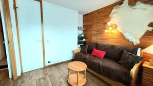 Una habitación acogedora con piso de madera, un sofá marrón con cojines rojos, una pequeña mesa de madera y una alfombra de piel blanca en la pared.