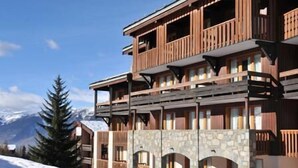 Un hotel a più piani con balconi in legno e pareti in pietra, sullo sfondo di una montagna innevata.