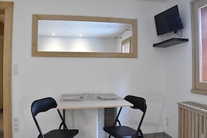 Un petit coin repas avec une table et deux chaises, un miroir au-dessus de la table, un téléviseur accroché au mur et une fenêtre sur la droite.